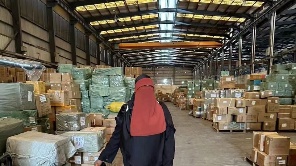 A woman in a veil walking through a large warehouse aisle lined with wrapped goods and pallets